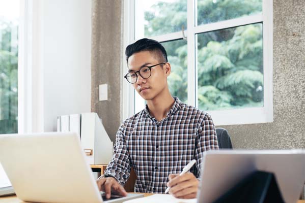Young serious Asian man in checked shirt and glasses reading information on laptop and making notes while sitting at table
